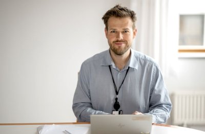 Mann i skjorte sitter ved et bord med en bærbar PC og smiler.