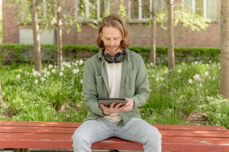 Mann med hodetelefoner studerer på nettbrett i en park.