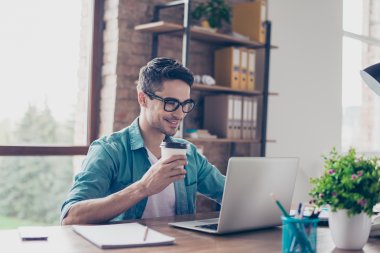 Mann med briller jobber på laptop og drikker kaffe ved skrivebord.