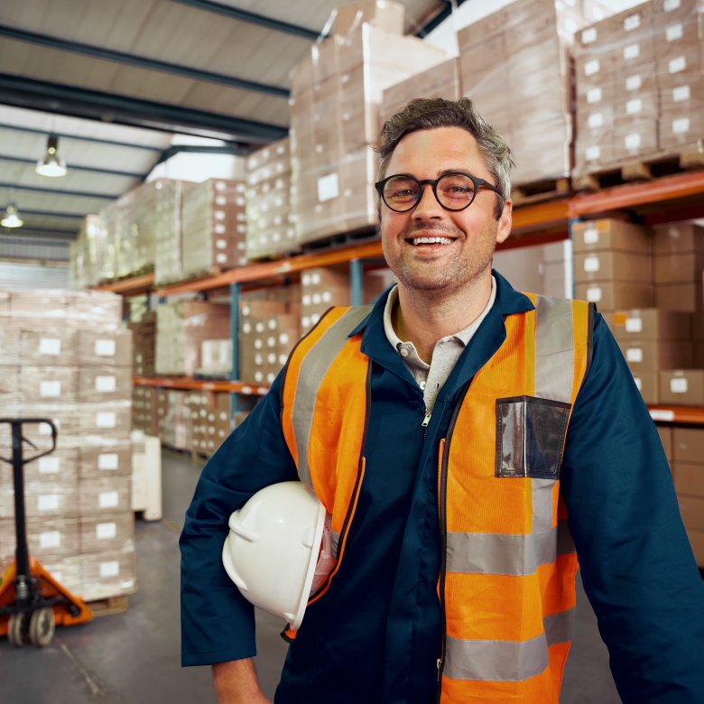 Mannlig arbeidsleder innen logistikk står med vernehjelm under armen i et varelager og smiler
