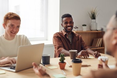 Tre personer smiler og diskuterer rundt et bord med bærbar PC og kaffekopper.