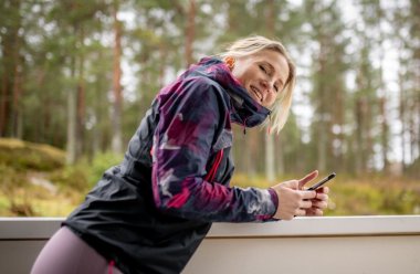 Kvinne i treningsklær smiler og holder en mobiltelefon utendørs i skogen.