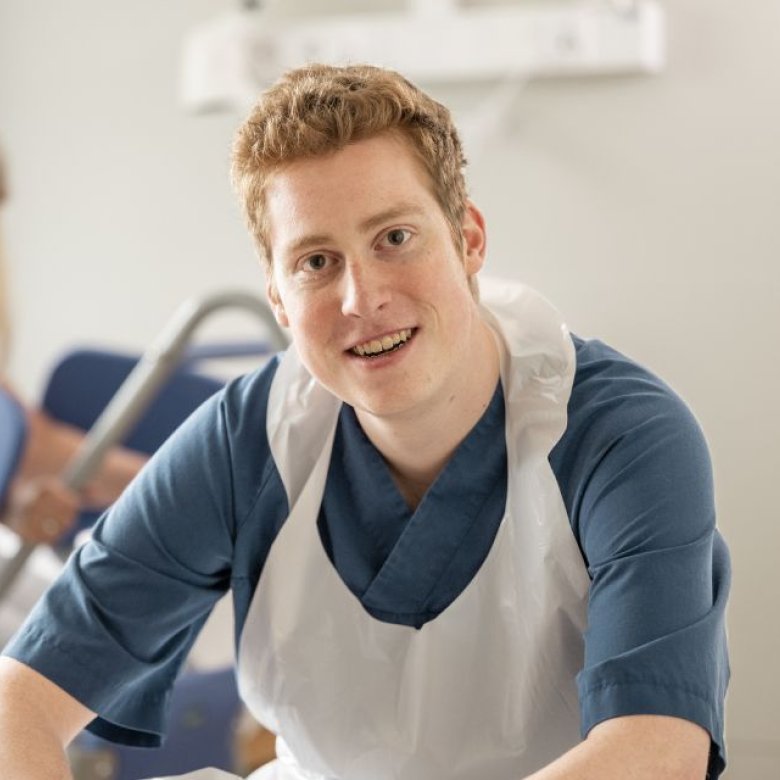 Bilde av en mannlig pleieassistent på jobb med en pasient i bakgrunnen 